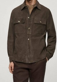 Uomo che indossa una giacca button-up scamosciata marrone scuro con tasche a patta sopra un maglione beige a coste e pantaloni marroni coordinati.