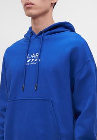 Homme portant un sweat à capuche bleu vif avec poche avant et texte blanc du logo "L/AB c/o Off-White" sur la poitrine, sur fond clair uni.