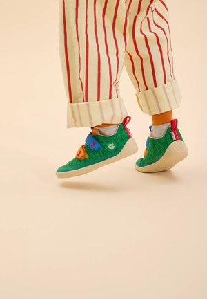 Grüne Schuhe mit einem strukturierten Obermaterial, ausgestattet mit blauen und orangefarbenen Klettverschlüssen. Weiße Gummisohlen, getragen mit gestreiften cremefarbenen und roten Hosen.