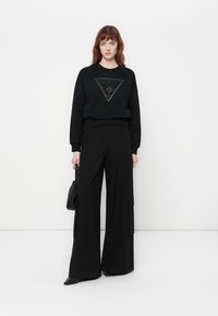 Sudadera negra con un logo triangular, combinada con pantalones negros de pierna ancha con bolsillos laterales. El modelo sostiene un pequeño bolso negro.