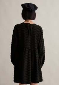 Schwarzes Kleid mit strukturiertem Streifenmuster, langen Ärmeln und gerafftem Bund. Verfügt über einen Bindeverschluss im Rücken und einen fließenden Rock.