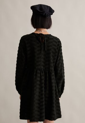 Schwarzes Kleid mit strukturiertem Streifenmuster, langen Ärmeln und gerafftem Bund. Verfügt über einen Bindeverschluss im Rücken und einen fließenden Rock.