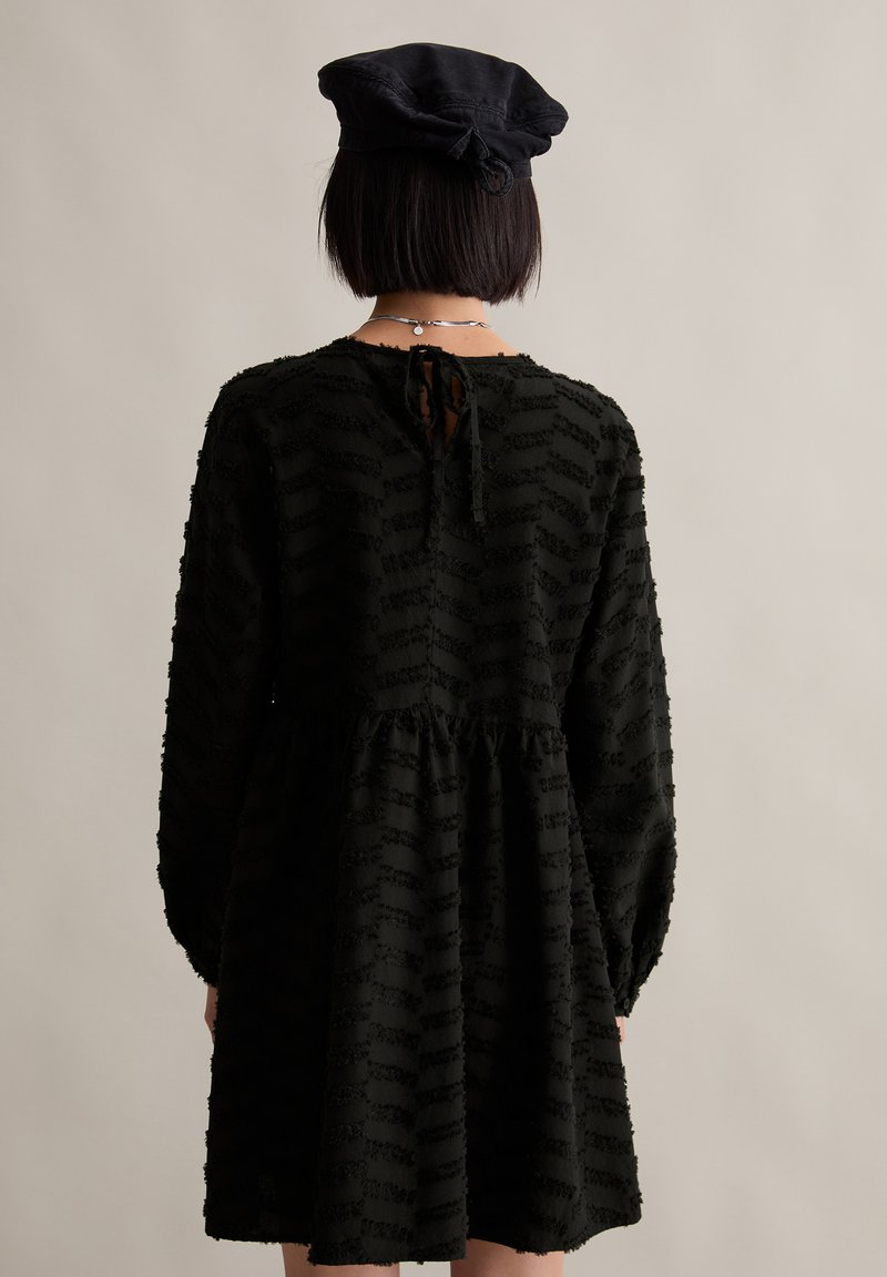 Schwarzes Kleid mit strukturiertem Streifenmuster, langen Ärmeln und gerafftem Bund. Verfügt über einen Bindeverschluss im Rücken und einen fließenden Rock.