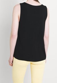 Osoba v bezrukávovém černém topu a světlých žlutých kalhotách, stojící zády k fotoaparátu na jednobarevném pozadí.