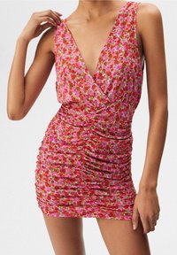 Mini-robe fleurie avec décolleté en V profond, tissu rose orné de motifs rouges et verts, détails froncés le long du corps, et design sans manches.