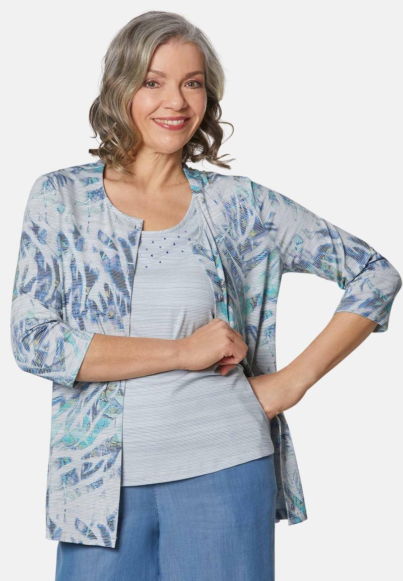 Lichte open-front cardigan in blauw en witte abstracte print, 3/4 mouwen, gecombineerd met een lichtgestreepte tanktop en blauwe broek.