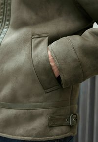 La chaqueta de ante verde oliva cuenta con un bolsillo lateral, un forro suave y una correa ajustable con detalle de hebilla en la cintura.