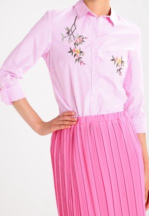 Kvinde iført en lyserød og hvid stribet skjorte med knapper og blomstret broderi samt en knaldpink plisseret nederdel, hånd på hoften.