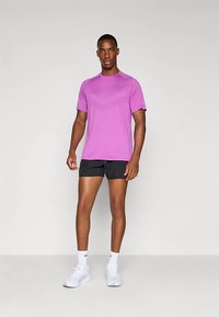 Hombre de pie vestido con una camiseta atlética corta de manga morada, pantalones cortos de correr negros, calcetines blancos y zapatillas blancas sobre un fondo liso.