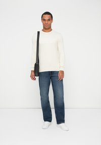 Suéter de algodón color crema claro con el logo "GUESS" en relieve, combinado con jeans rectos de color azul oscuro y zapatillas blancas, complementado con un bolso negro.