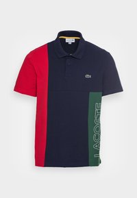 Polo avec des blocs de couleur rouge, bleu marine et vert, à manches courtes, et un logo vert. Fabriqué en coton, avec un col standard et des boutons.