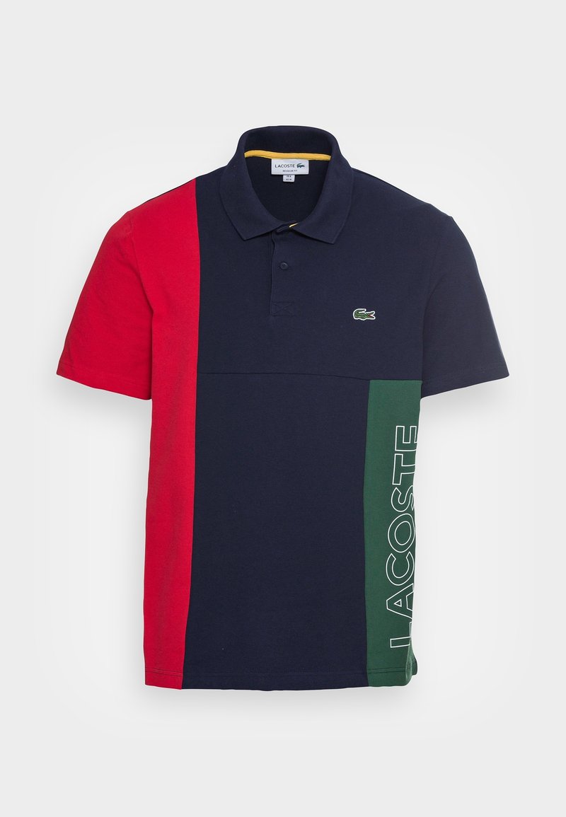 Polo avec des blocs de couleur rouge, bleu marine et vert, à manches courtes, et un logo vert. Fabriqué en coton, avec un col standard et des boutons.