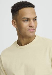 Lys beige bomulds t-shirt med rund hals, der har en glat tekstur og minimal syning. Modellen har kort, krøllet hår.