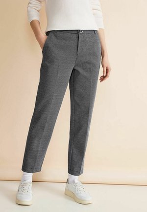 Personne portant un pantalon gris texturé à longueur de cheville, des baskets blanches, des chaussettes blanches et un pull blanc côtelé, les mains détendues le long du corps.