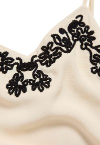 Cremefarvet cami-top med sort broderet blomsterdesign ved halsen. Blød stof med en glat tekstur, udstyret med tynde justerbare stropper.