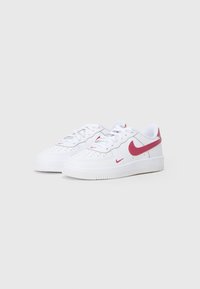 Baltas zemas Nike sporta apavi ar sarkanu Swoosh logotipu, šņorēm un caurumiņiem uz pirkstu zonas, izvietoti uz neitrālas gaišas fona.