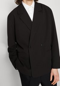 Blazer nero doppiopetto con rever a punta, due bottoni e tasche frontali, indossato sopra una camicia bianca con colletto.