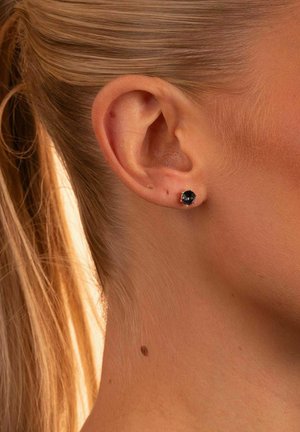 Svarta stensetter i guld-tonad metall, med rund form och klorinställning, visade på en modells öra mot blonda hår.