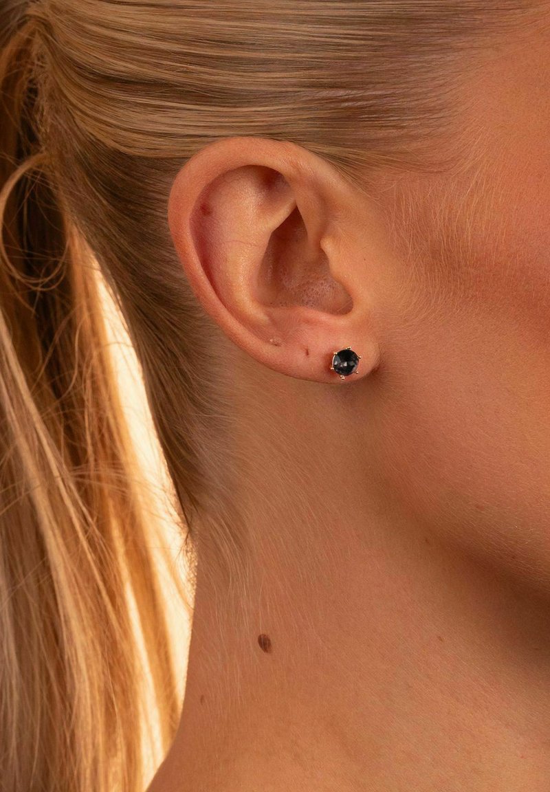 Schwarze Steinstud-Ohrringe aus goldfarbigem Metall, mit runder Form und Krappenfassung, präsentiert am Ohr eines Modells mit blonden Haaren.