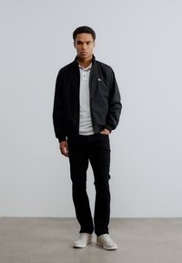 Μαύρο ελαφρύ μπουφάν bomber πάνω από ένα λευκό πόλο, σε συνδυασμό με μαύρο παντελόνι και λευκές αθλητικές παπούτσια. Απλός, καθαρός σχεδιασμός χωρίς μοτίβα.