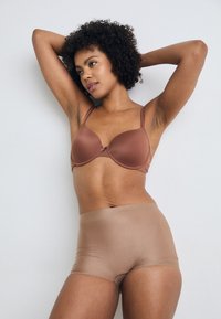 Bra in Braun mit geformten Cups und verstellbaren Trägern, kombiniert mit nahtlosen, braunen, hochgeschnittenen Shorts. Glatte Textur, minimale Hardware-Details.