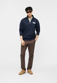 Homme portant des lunettes de soleil noires, un sweat-shirt bleu marine à demi-fermeture éclair avec le logo "GAP", un polo vert clair, un pantalon marron et des baskets couleur tan, debout les mains dans les poches.