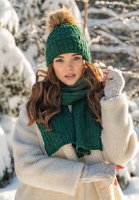 Gorro de punto verde con un pompón de piel sintética, bufanda de canalé a juego y abrigo de forro polar crema. Textura suave con patrones detallados.