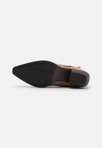 Zapato de ante marrón con diseño de punta afilada, superficie texturizada y suela de goma negra. Incluye un pequeño tacón apilado para mayor soporte.