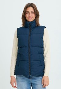 Tums zils pūkains aizdaris ar augstu apkakli, priekšējo rāvējslēdzēju un divām sānu kabatām; ar šuvēm horizontālās daļās, lai radītu tekstūru.