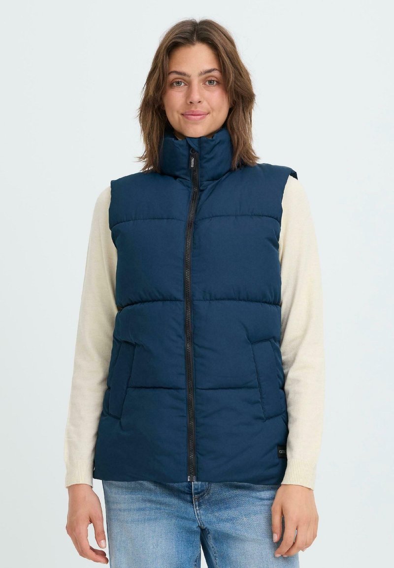 Tums zils pūkains aizdaris ar augstu apkakli, priekšējo rāvējslēdzēju un divām sānu kabatām; ar šuvēm horizontālās daļās, lai radītu tekstūru.