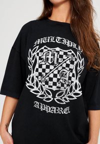 Camiseta negra de gran tamaño con un diseño gráfico en blanco desgastado. El diseño incluye un patrón de cuadros y acentos de laurel. Mangas cortas.