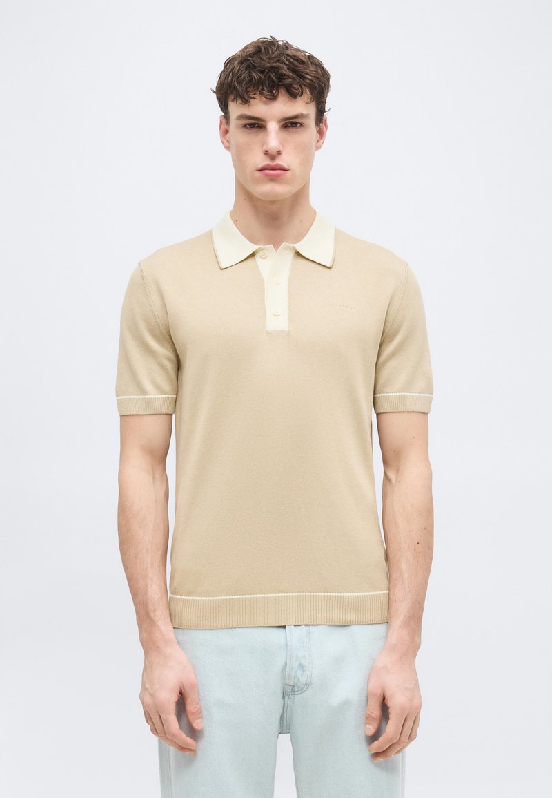 Jeune homme portant un polo beige à manches courtes avec un col blanc et un jean bleu clair, debout devant un fond blanc uni.