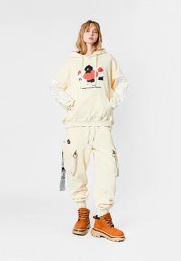 Joven con sudadera crema con un gráfico, pantalones cargo a juego y botas de cordones color tostado, de pie contra un fondo blanco liso.