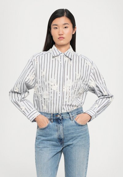 Bela in siva črtasta srajca z dolgimi rokavi in klasičnim ovratnikom, okrašena s cvetličnim vezenjem, v kombinaciji z visokim pasom modrimi kavbojkami.