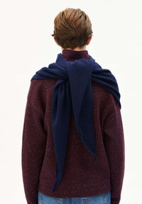 Persoon van achteren gezien, gekleed in een kastanjebruine gespikkelde trui en een blauwe gebreide sjaal die over de schouders hangt, gecombineerd met blauwe spijkerbroek.