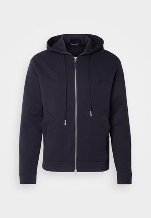 Tamno plavi zip kaput od mješavine pamuka. Sadrži kapuljaču s vezicom, prednje džepove i suptilni otisnuti logo na prsima.