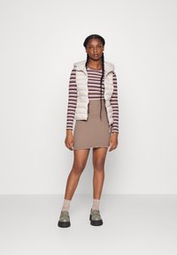 Colete puffy bege claro sobre uma blusa de mangas longas em marrom e creme às riscas, combinado com uma saia texturizada castanha e sapatos de plataforma verde-oliva.