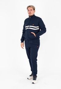 Tuta navy in misto cotone; giacca con zip e strisce bianche, tasche frontali; pantaloni abbinati con vita elastica; sneakers bianche.