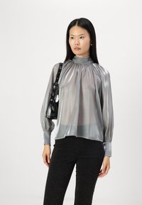 Blusa grigia trasparente con colletto alto, maniche a sbuffo e dettagli arricciati; abbinata a pantaloni neri glitterati e una piccola borsa nera.