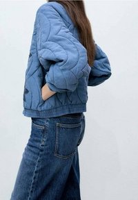 Niebieska pikowana kurtka z oversize'owymi rękawami i teksturowanym wzorem, w połączeniu ze spodniami dżinsowymi o średnim praniu, sfotografowana z boku.