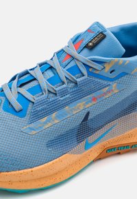 Blå atletiksko med et tekstureret mesh-øvre, GORE-TEX-mærke og en svampet, orange mellemsål med akvagrønne accenter og snørebånd.