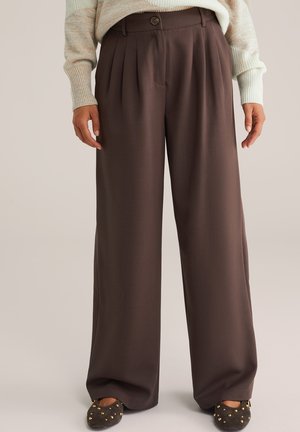Person trägt hoch taillierte, weit geschnittene braune Hosen kombiniert mit einem hellbeigen Strickpullover und dunkelbraunen, mit Nieten besetzten Schuhen.
