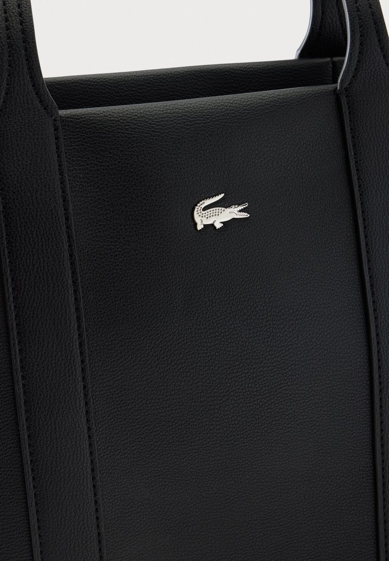 Borsa in pelle nera testurizzata con un emblema logo coccodrillo argento centrato sul pannello frontale e doppi manici visibili nella parte superiore.