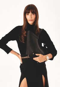 Borsa in pelle nera con una forma strutturata, logo impresso e dettagli in oro, tenuta da una persona che indossa un top nero e una gonna con spacco laterale.