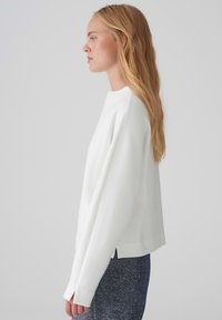 White sweatshirt with a high neckline, long sleeves, and a loose fit. The hem has side slits. Worn with textured dark blue trousers.
