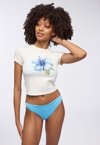 T-shirt corta di colore crema con stampa di fiore di ibisco blu; abbinata a pantaloni bikini azzurri. Tessuto liscio, maniche corte, design aderente.