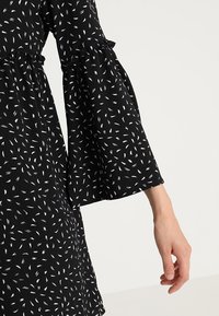 Vestido negro con patrones en forma de hoja blanca, con mangas acampanadas y una cintura con volantes, hecho de tela ligera.