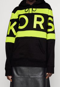 Sudadera negra de material suave, con atrevidas rayas verde neón y la palabra "KORS" en letras. Incluye capucha con cordón y bolsillo frontal.