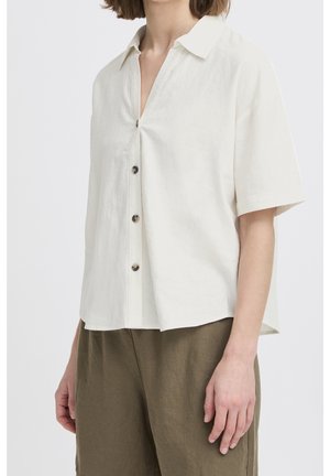 Femme portant une chemise blanche à manches courtes et ample, boutonnée, et un pantalon marron taille haute sur un fond uni.