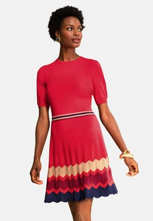 Femeie purtând o rochie roșie cu mâneci scurte, cu o talie cu dungi și o fustă cu model șerpuiri în bej, roșu și bleumarin, accesorizată cu brățări și cercei.